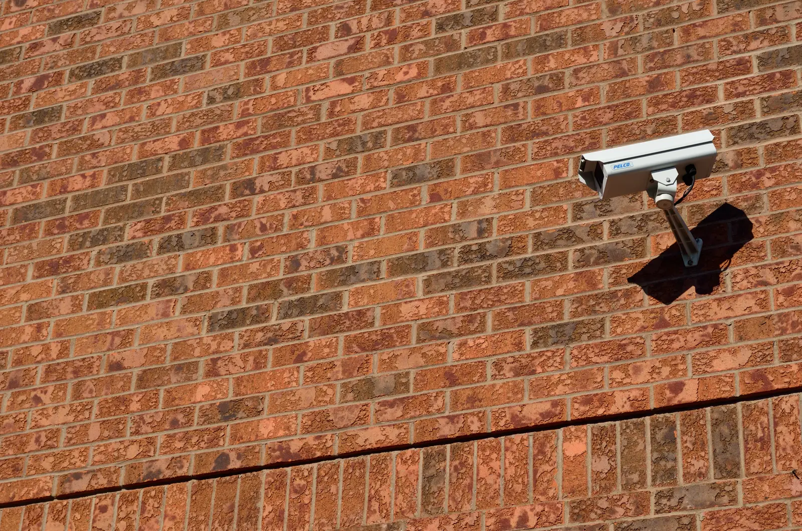A white security camera on a metal arm mounted high on a sunlit red brick wall, casting a shadow beside it