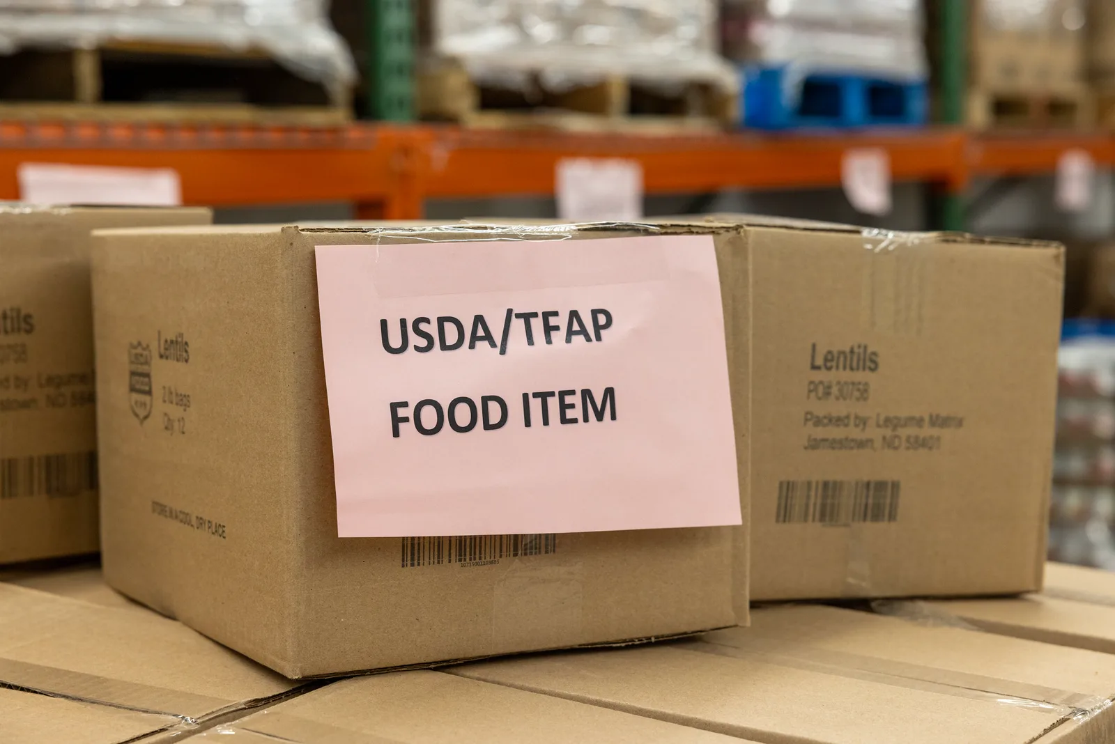 Cardboard cases of lentils, one with a pink sign reading USDA/TFAP Food Item, stacked in front of orange warehouse racks holding wrapped pallets