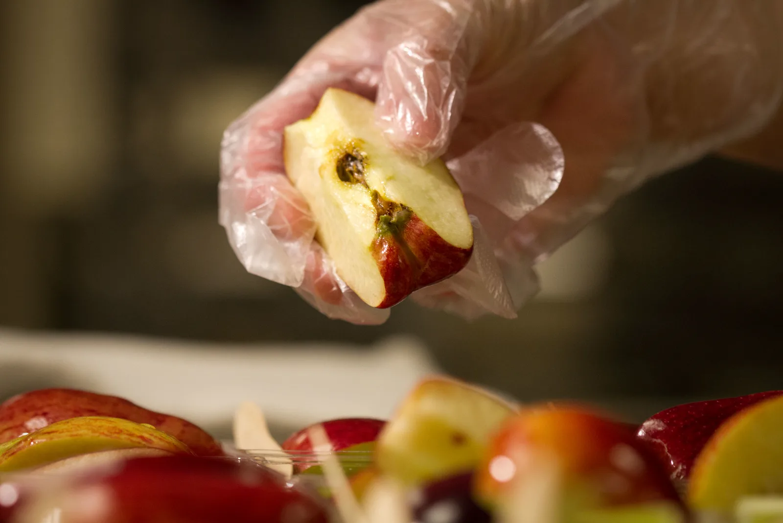 A hand in a clear plastic glove holds a wedge of red apple above a tray of cut apple slices