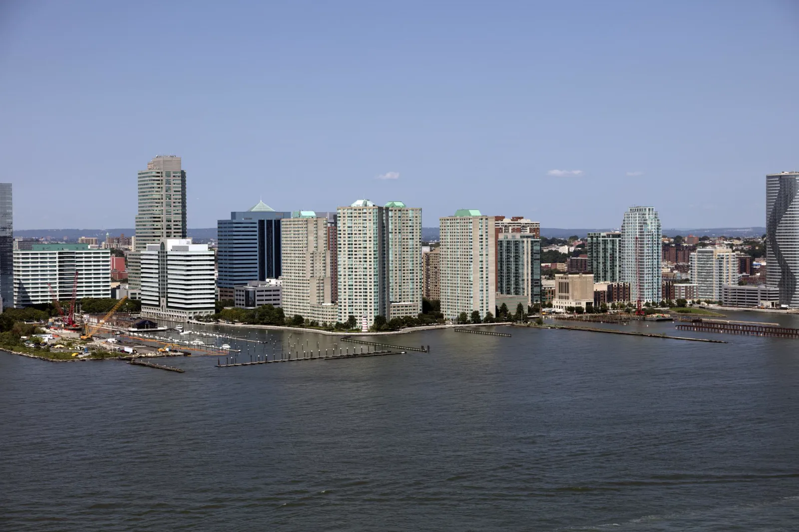 Apartment and office towers stand along a waterfront with a marina, piers and a small striped lighthouse, seen across a wide river under a clear blue sky