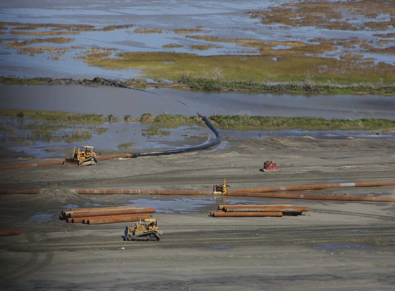 Two bulldozers and long rust colored pipes on a wide stretch of new sand beside channels of water and green marsh grass