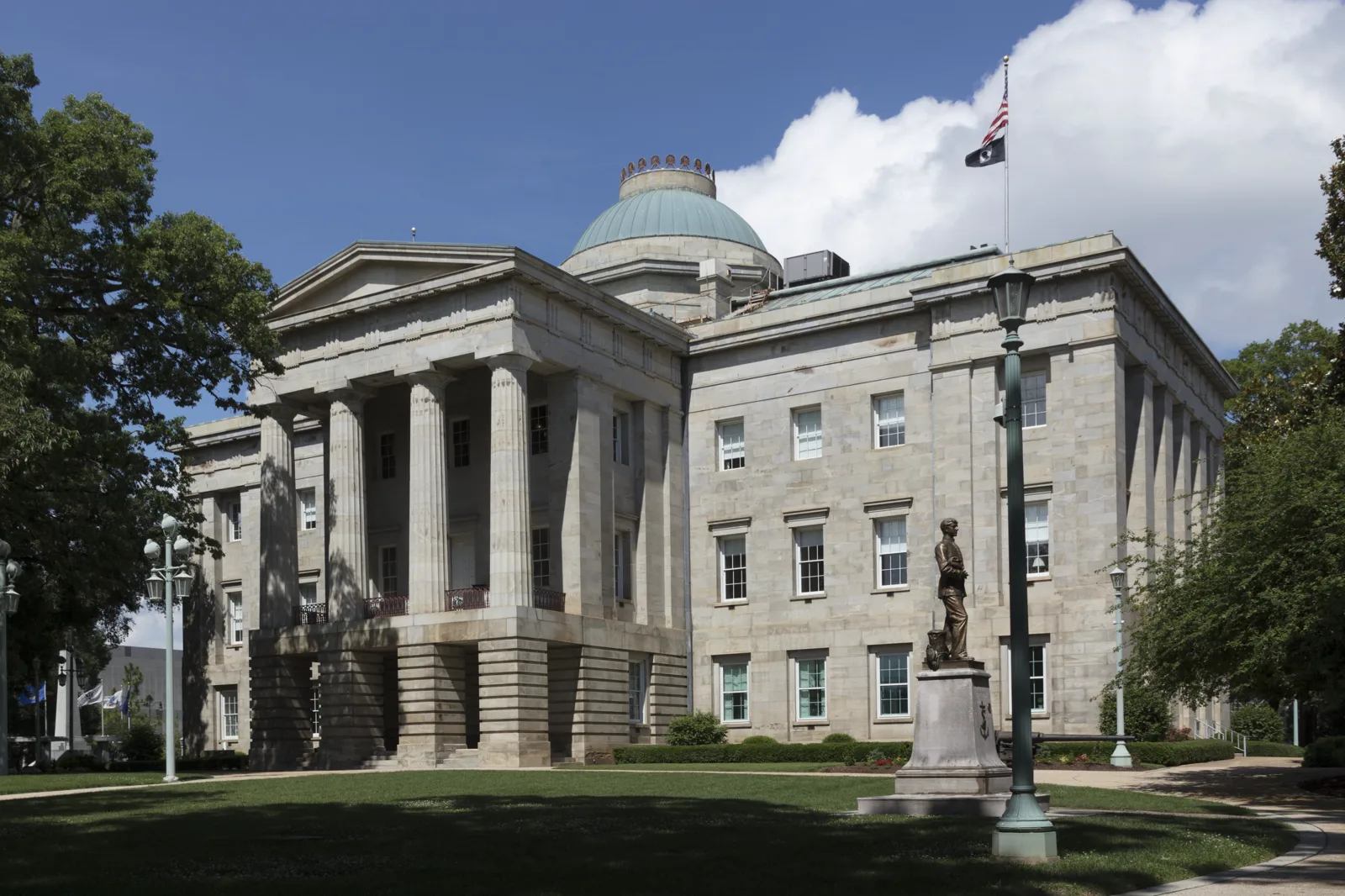 A pale stone capitol building with a columned portico and a green copper dome, a bronze statue on a stone pedestal and a lamppost on the lawn in front, and an American flag flying from the roof under a blue sky