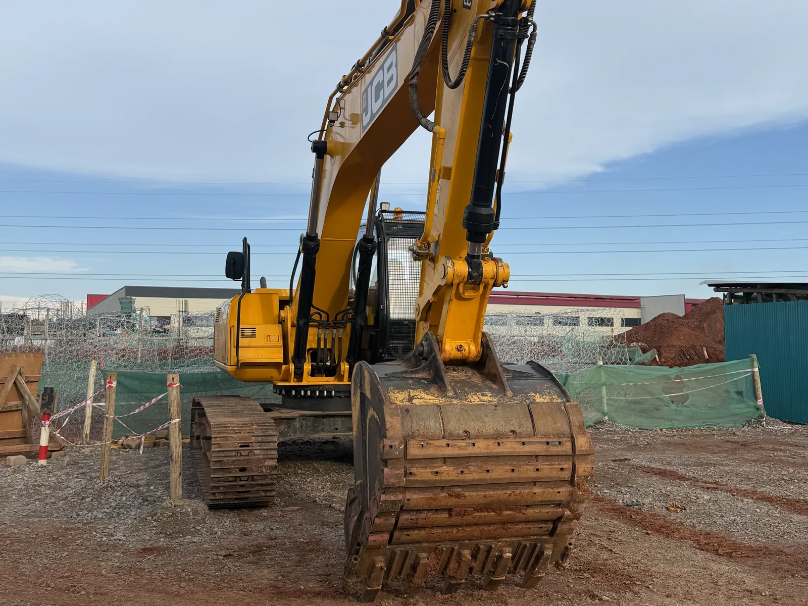 A yellow excavator with its bucket resting on bare dirt at a fenced construction site, with low industrial buildings and a mound of red soil behind it under a cloudy sky