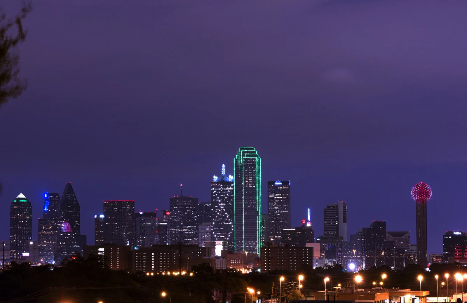 The downtown Dallas skyline at dusk under a purple sky, with one tower outlined in green light, a spherical tower lit in red on the right, and street lights along a dark tree line in the foreground