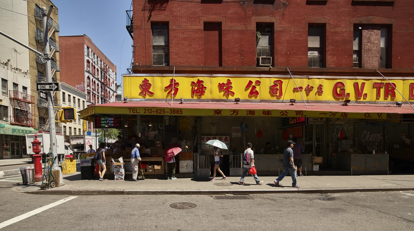 A grocery and seafood store under a long yellow sign with red Chinese characters, its awning shading produce stands, as a few shoppers, two under umbrellas, walk past on a sunny sidewalk below a red brick building