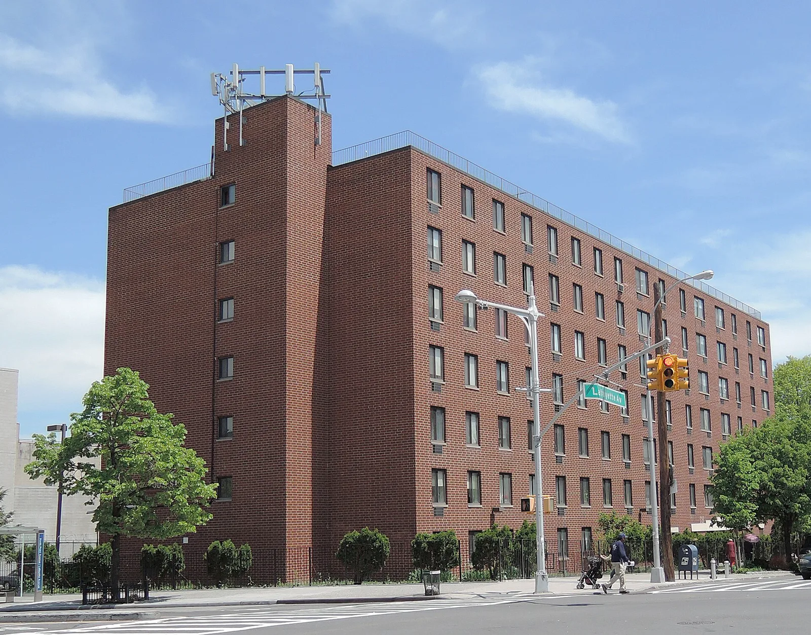 A seven-story red brick apartment building on a street corner under a blue sky, with a traffic light, a street sign and a person pushing a stroller in the distance