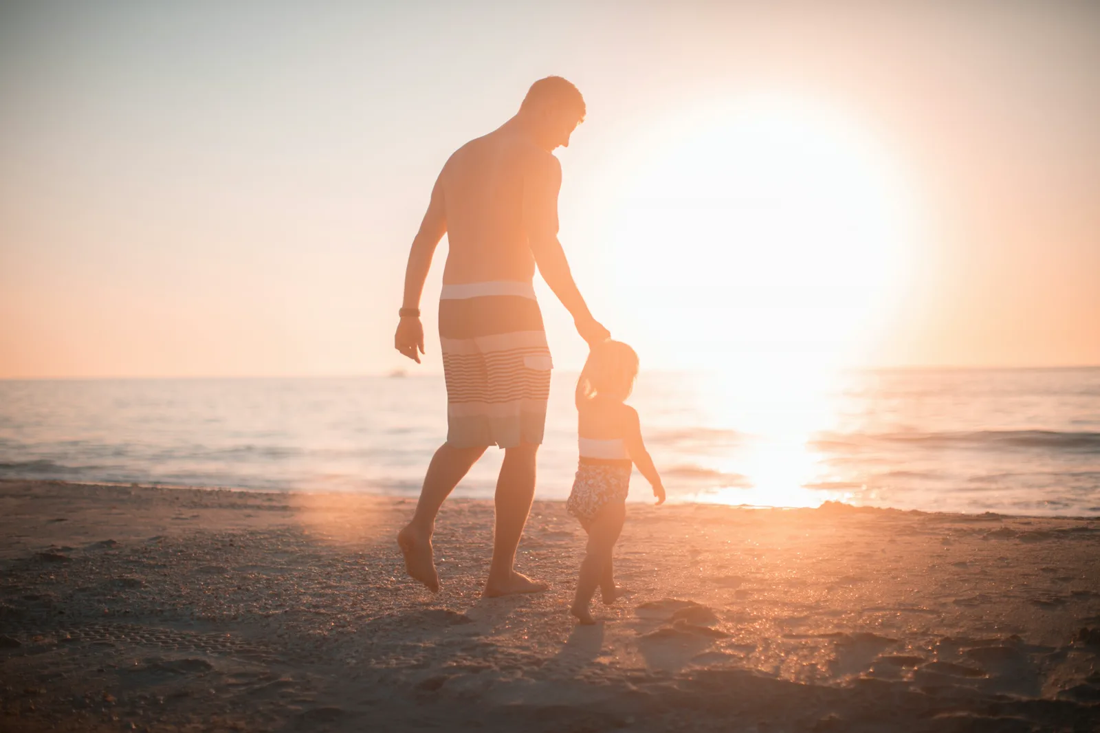A man and a small child, both in silhouette against a low sun, walk hand in hand along a beach at the water's edge