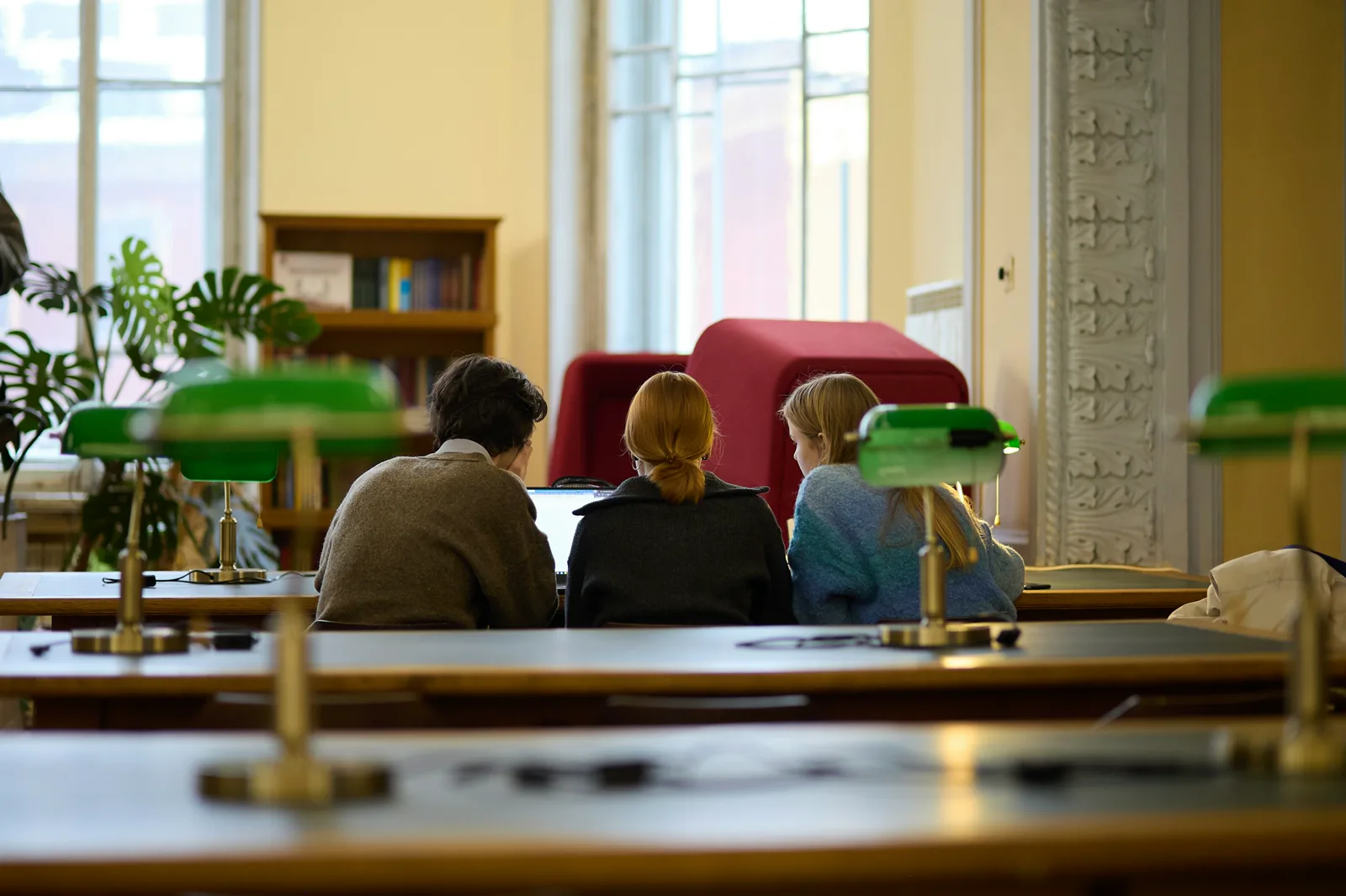 Three people seen from behind sit side by side at a long wooden desk looking at a laptop in a library reading room with green desk lamps, tall windows and a bookcase