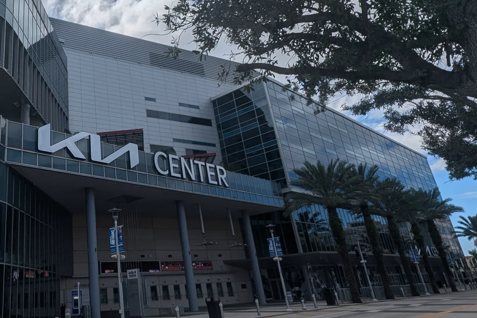 The glass and silver front of the Kia Center arena, with the Kia Center name over the entrance, a row of palm trees, and the branches of a large oak across a cloudy sky