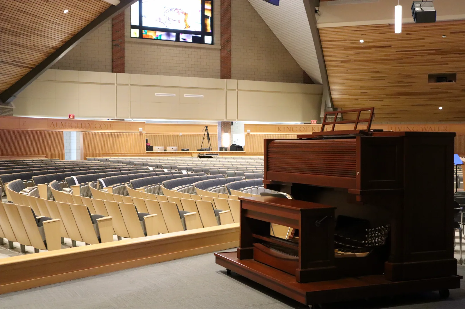 Rows of empty seats in a large church auditorium with wood paneled walls and ceiling, a stained glass window high on the far wall and an organ console in the foreground