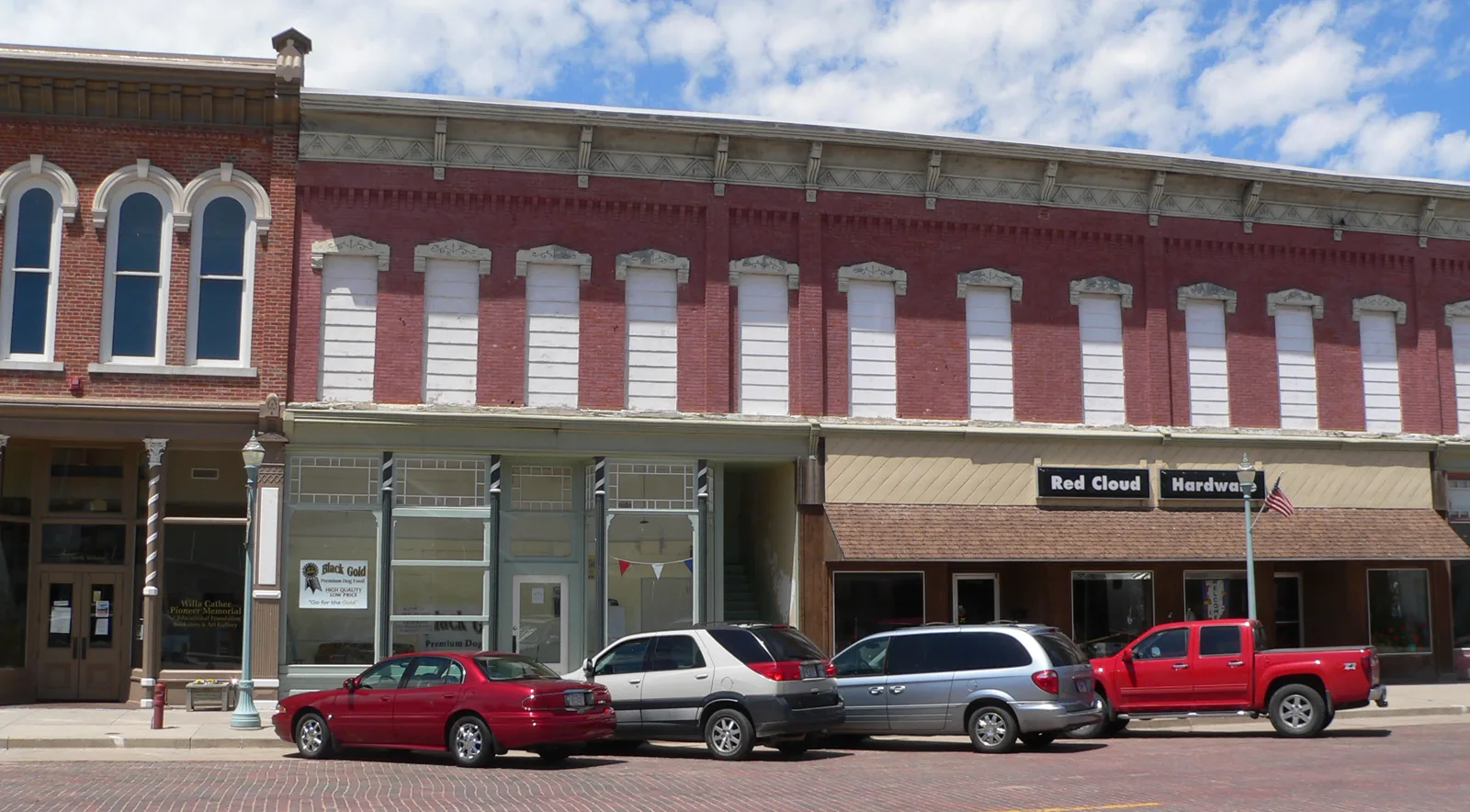A two-story red brick commercial block from the 1880s with ornate window hoods and its upper windows boarded with white panels, a hardware store and an empty storefront at street level, and cars parked on a brick-paved street