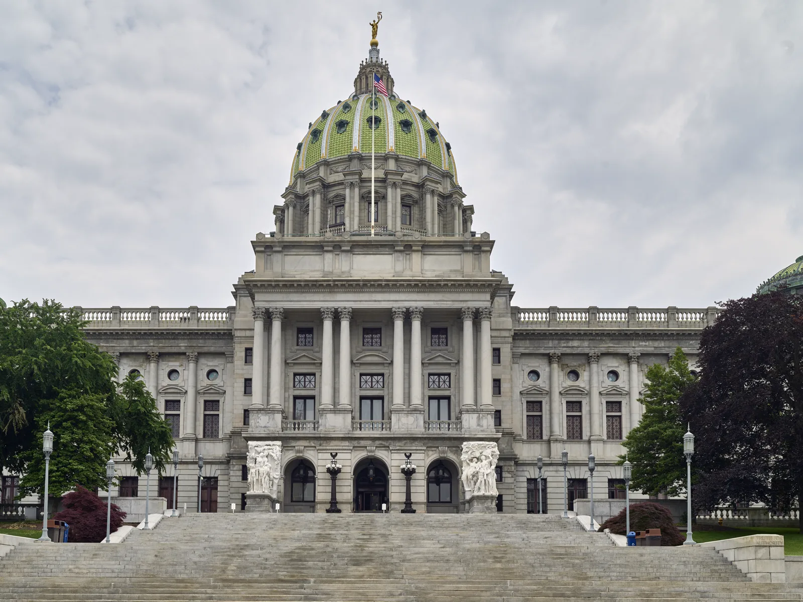 A granite capitol building with a green tiled dome topped by a gilded statue, a columned portico and two white sculpture groups beside the entrance, above a wide flight of stone steps under a cloudy sky