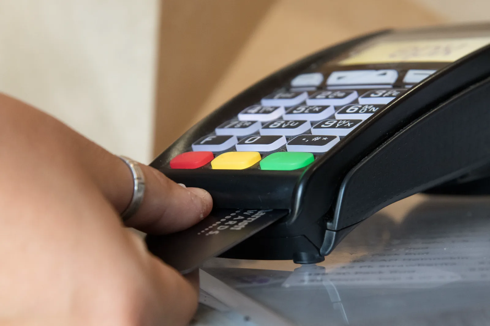 A close view of a hand with a silver ring inserting a payment card into a black card terminal with a numeric keypad and red, yellow and green keys