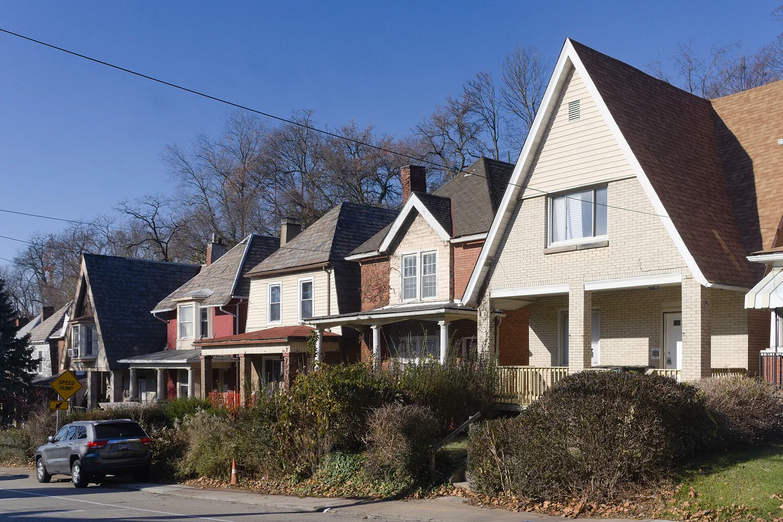 A row of brick and sided houses with steep gabled roofs along a sloping street under a clear blue sky, with bare trees and a parked car