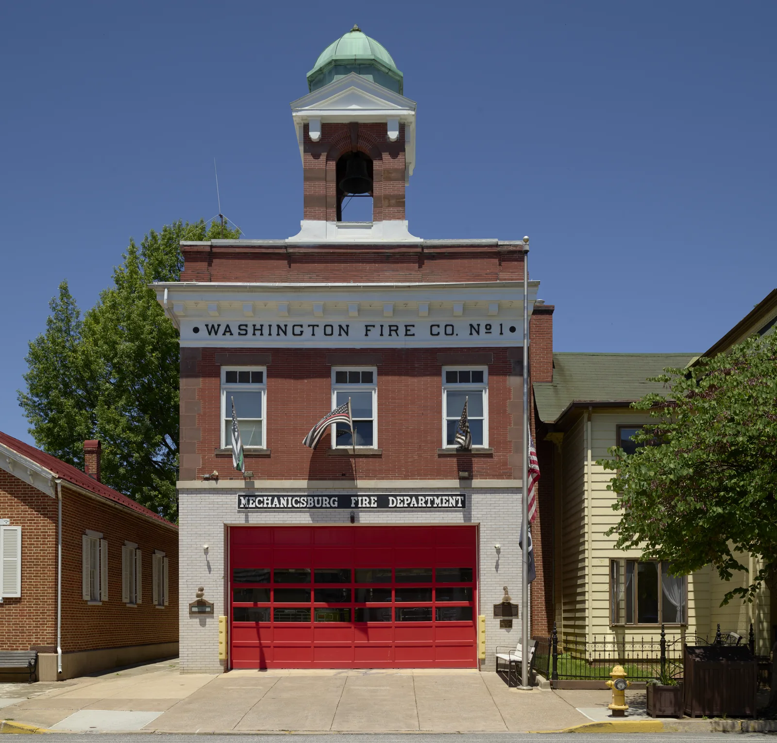 A two-story red brick firehouse with a green domed bell tower, a sign reading Washington Fire Co. No. 1 and Mechanicsburg Fire Department, and a red garage door on an empty street under a blue sky