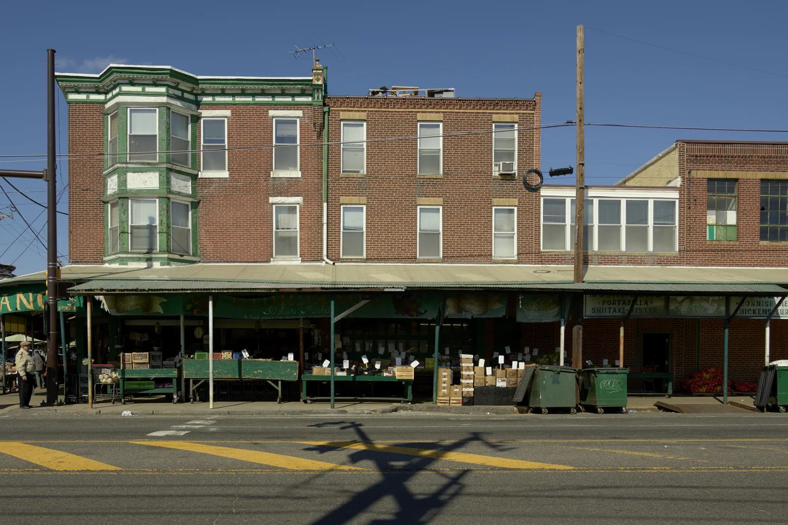Produce stands under a green metal awning along a row of brick buildings, with crates of vegetables, two green dumpsters at the curb and an empty street in the foreground