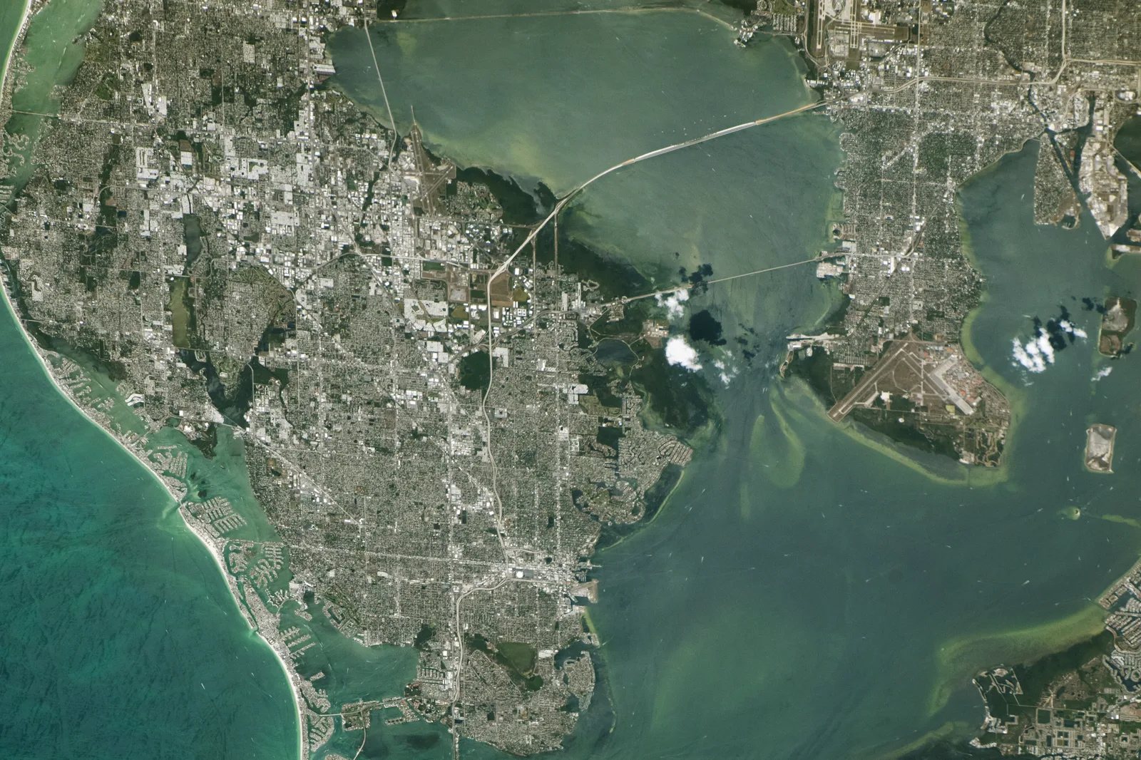 A view from space of a densely built peninsula between the turquoise Gulf of Mexico and the green waters of a wide bay, with long bridges crossing the bay to another city and an airfield on a point of land at right