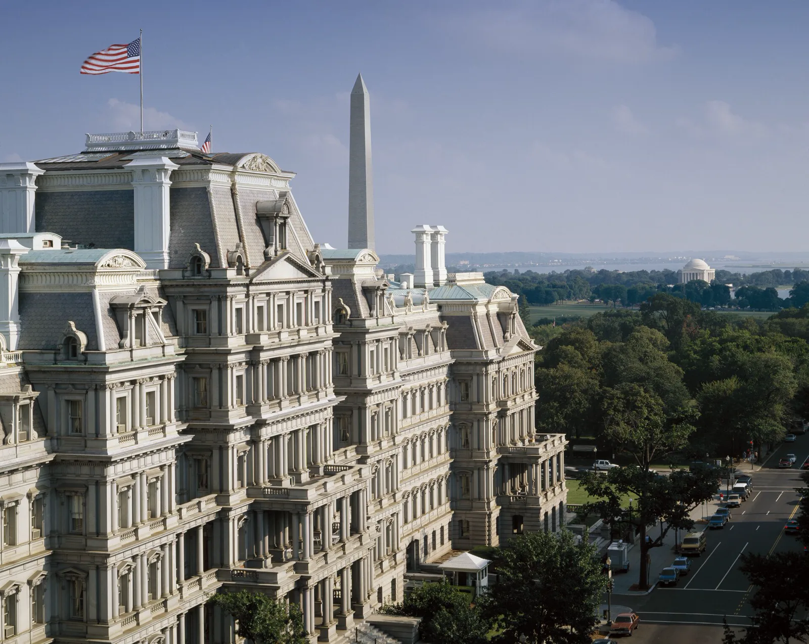 A large gray granite building with a mansard roof, rows of columns and an American flag on top, seen from above, with the Washington Monument and a domed memorial beyond the trees and a street with parked cars on the right