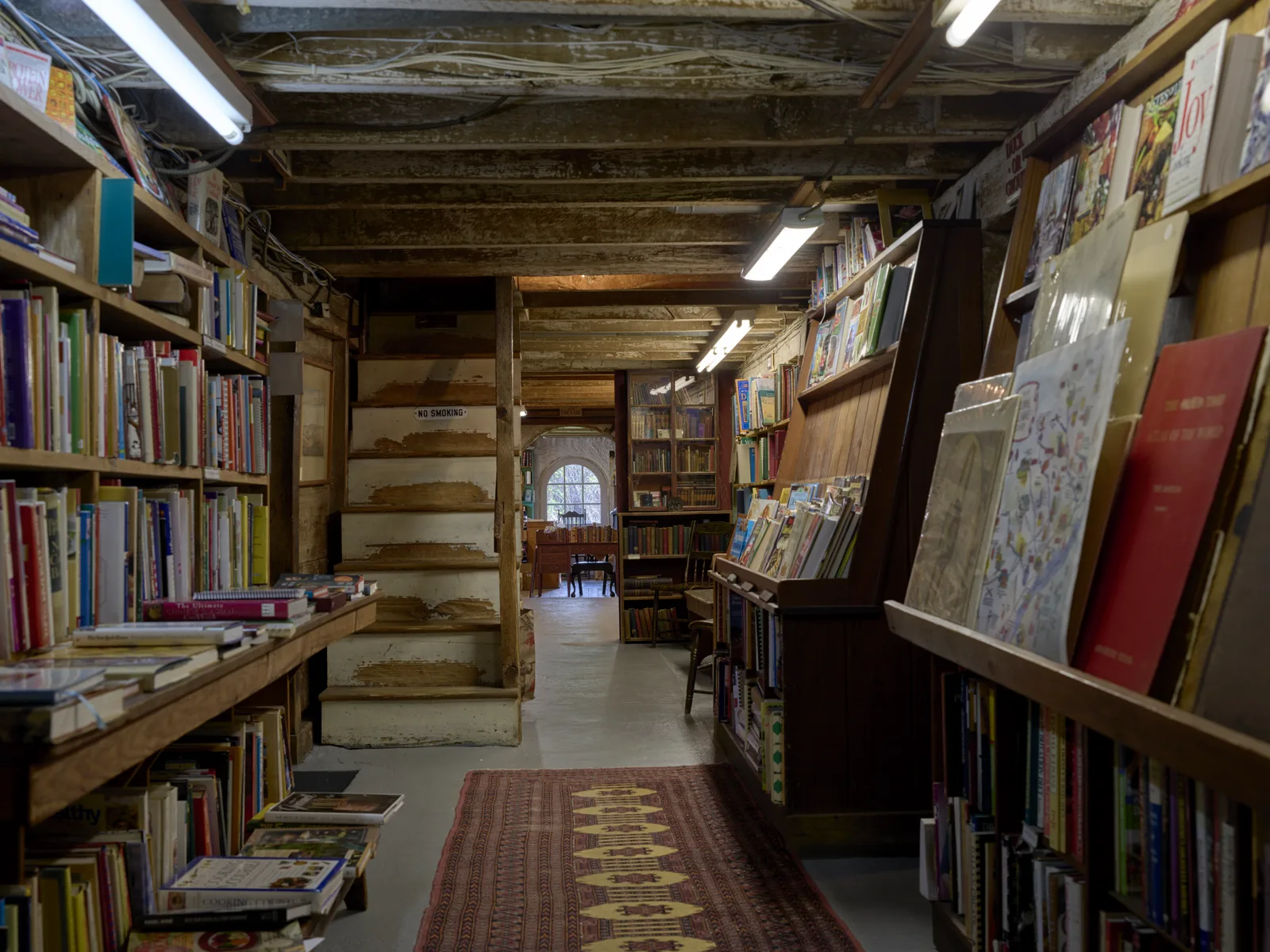A narrow aisle between wooden shelves packed with books, under a low ceiling of old beams and wiring, with a patterned runner on a concrete floor and an arched window at the far end