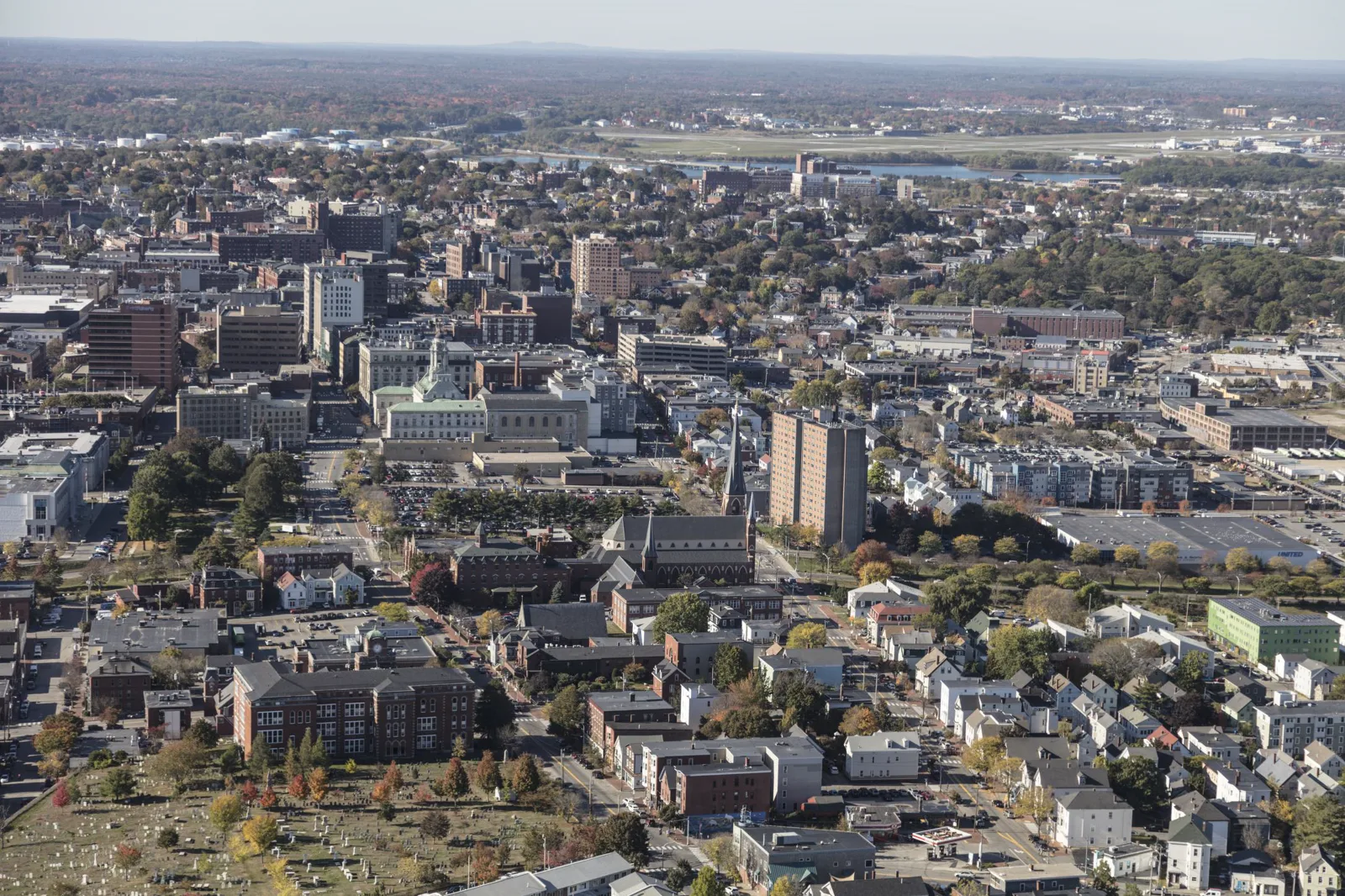 Aerial view of downtown Portland, Maine, in autumn: brick buildings, church steeples, a tall apartment tower and rows of houses, with a river and an airport in the distance