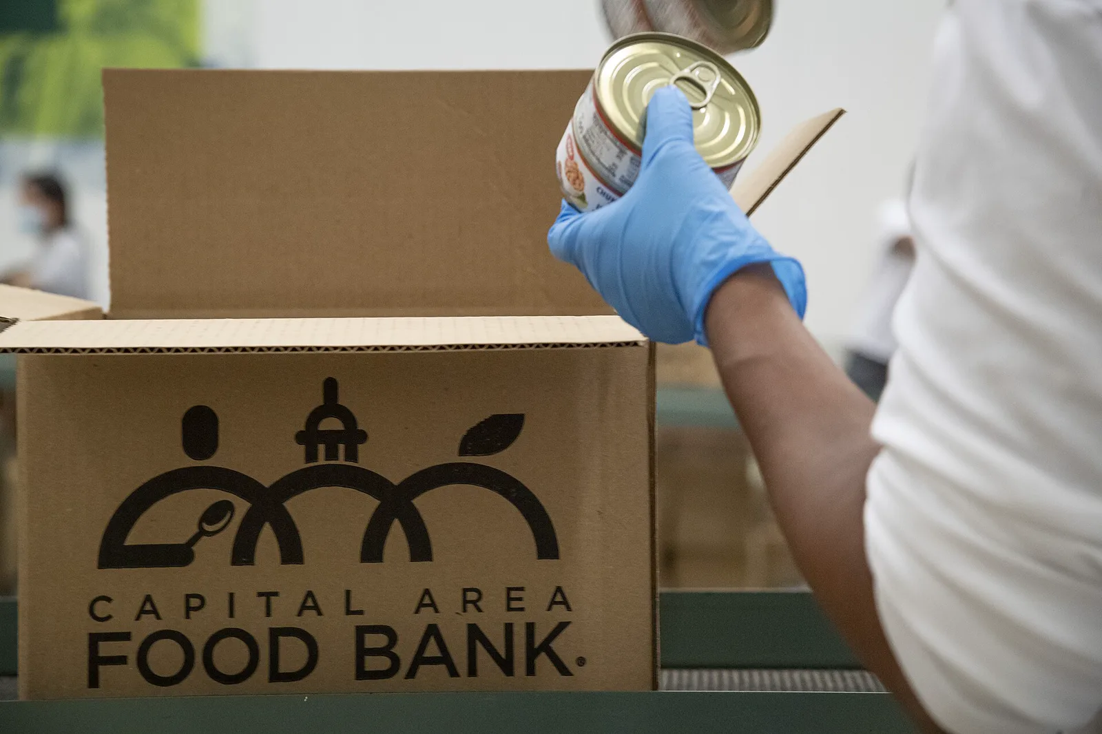A gloved hand holds a can of food over an open cardboard box printed with the Capital Area Food Bank logo on a packing line