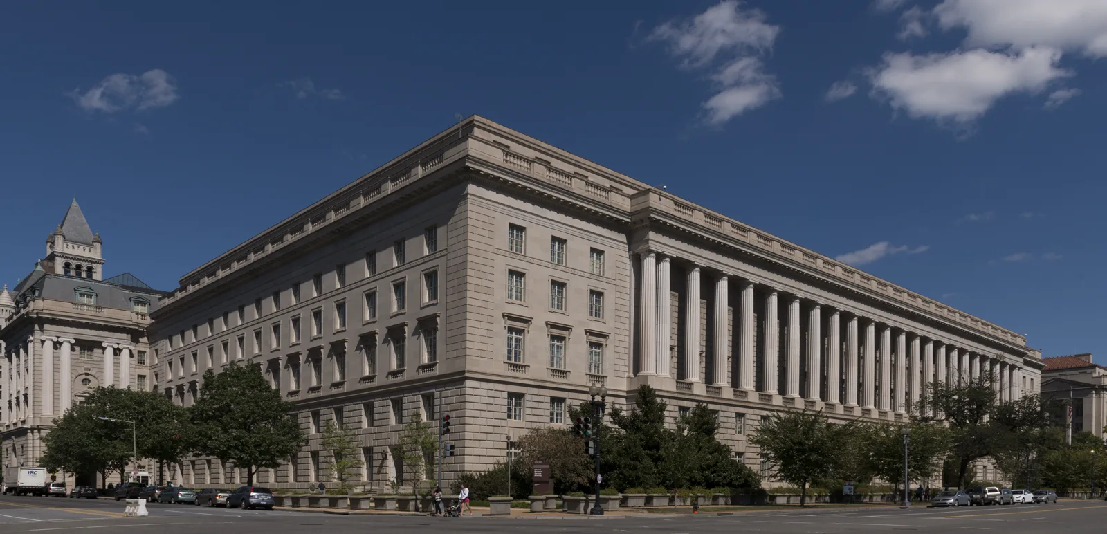 A long stone government building with a row of tall columns along one side, seen from a street corner under a blue sky with trees along the sidewalk