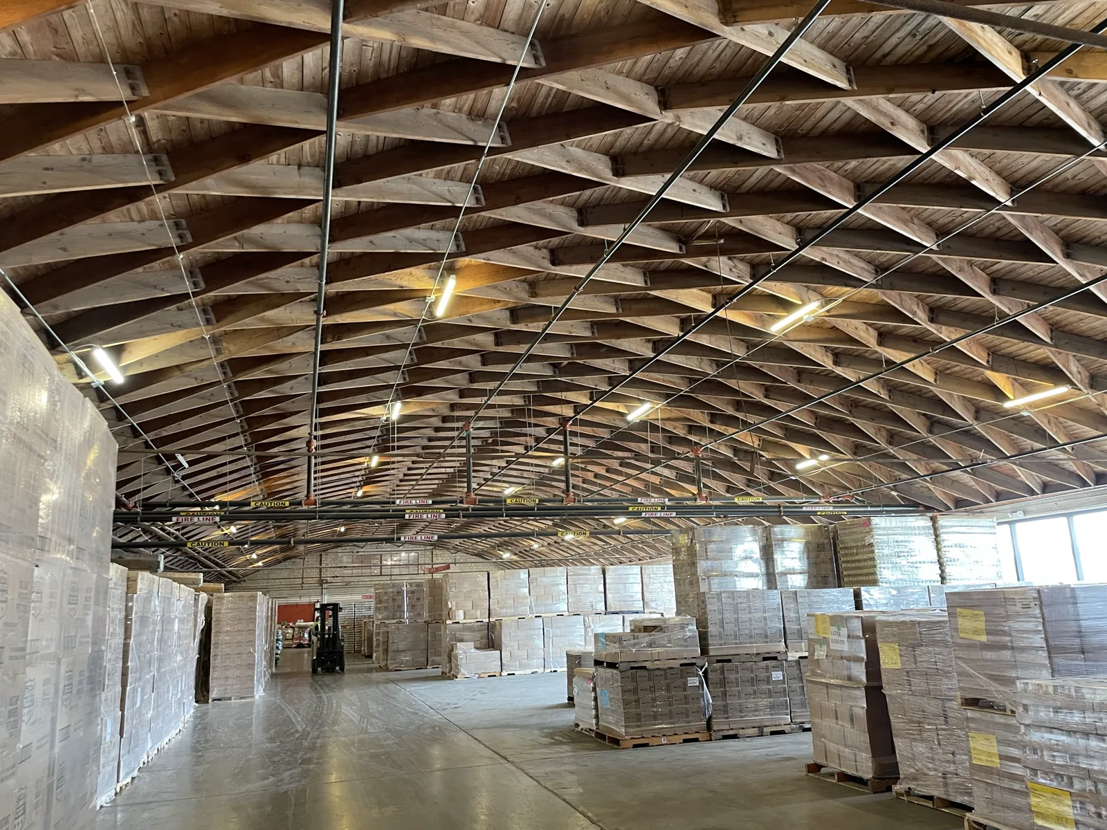 Rows of shrink-wrapped pallets of boxed food stand on the concrete floor of a large warehouse under a wooden truss roof, with a forklift in the distance