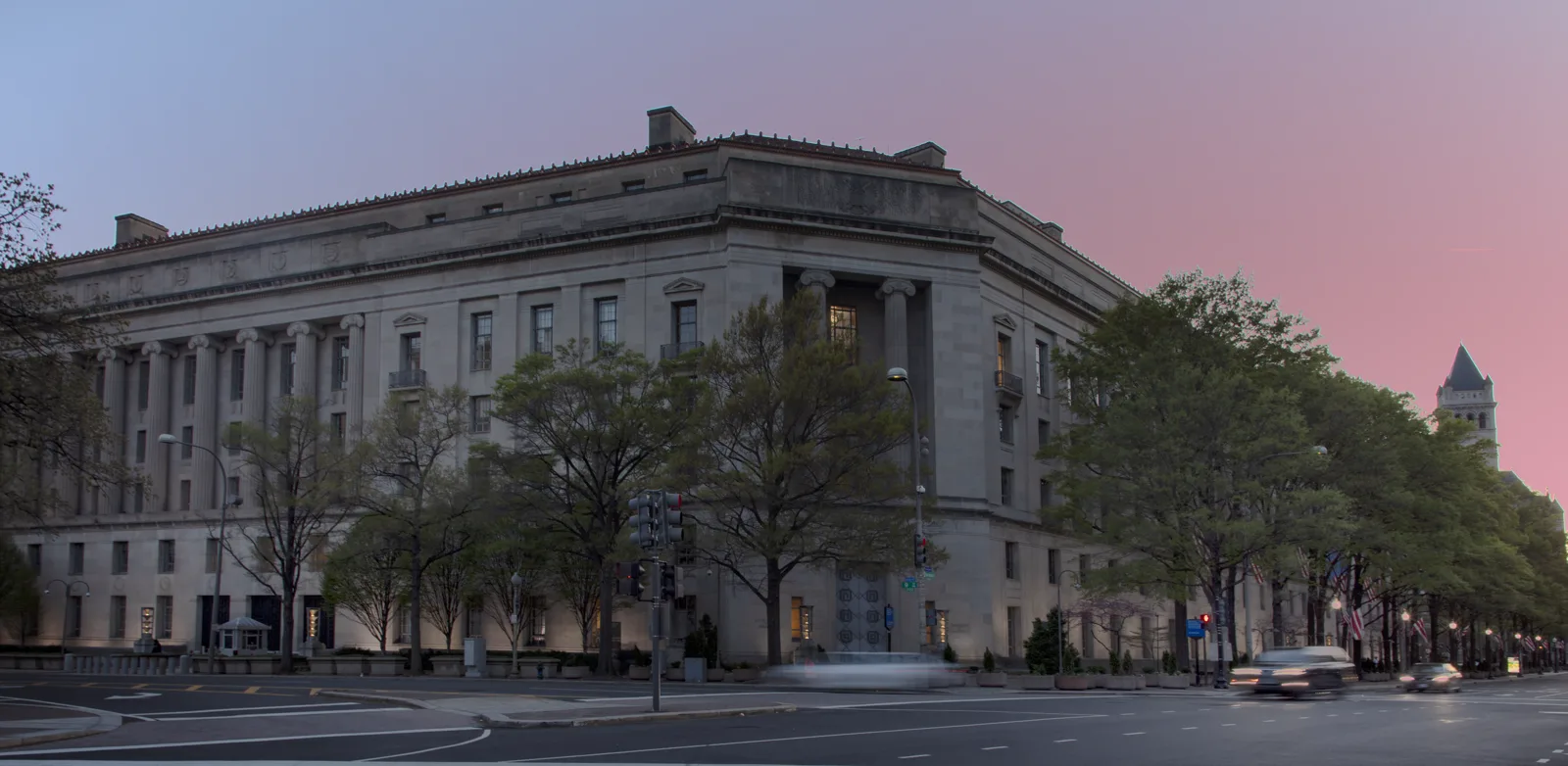 A large gray stone government building with tall columns and rows of lit windows at dusk, trees along the sidewalk, traffic lights and blurred cars on the street in front, under a pink and blue sky