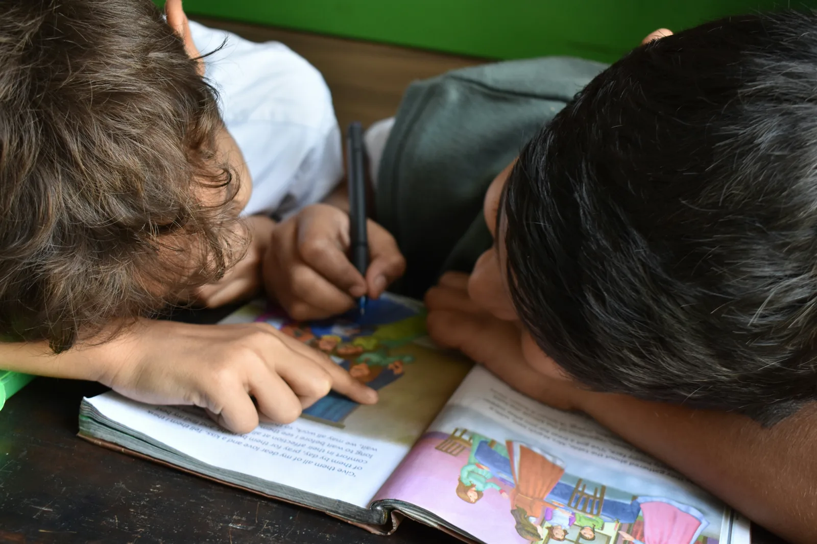 Two children seen from above lean their heads over an open picture book on a wooden table, one holding a pen and the other pointing at the page