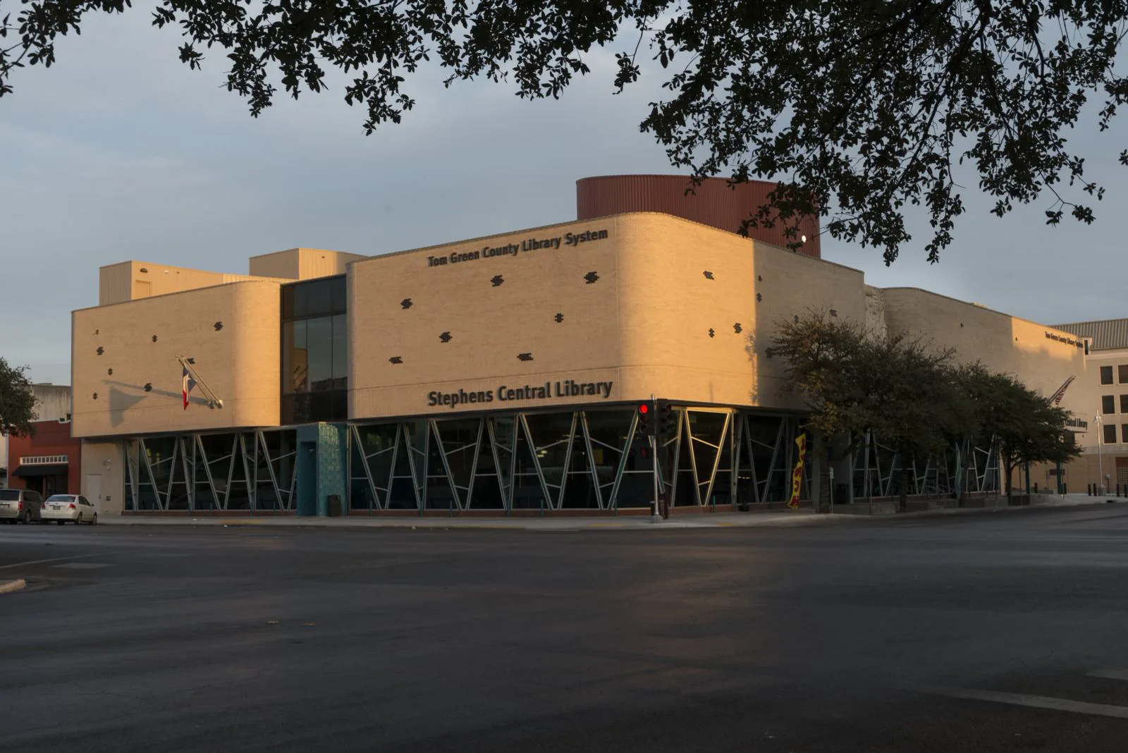 A cream brick library building with rounded corners and a wall of angled glass windows at a street corner, lettered Stephens Central Library and Tom Green County Library System, lit by low sun under overhanging tree branches