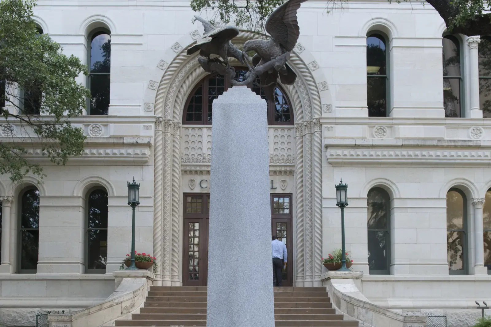 A pale stone building with arched windows and a carved arched entrance at the top of wide steps, with a granite obelisk topped by bronze eagles standing in front of it