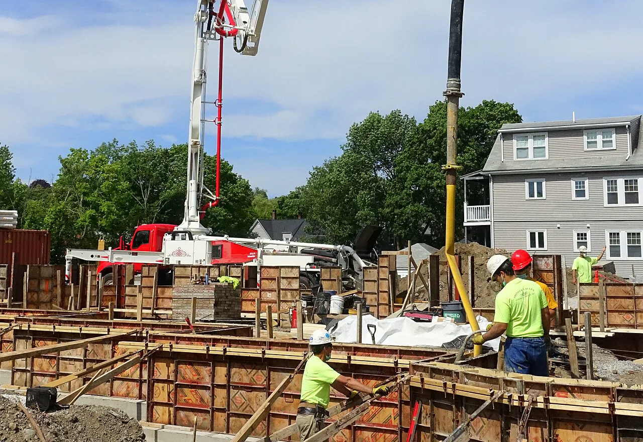 Workers in hard hats and bright yellow shirts stand among wooden foundation forms while the boom of a red and white concrete pump truck lowers a hose over them, with trees and a gray house behind