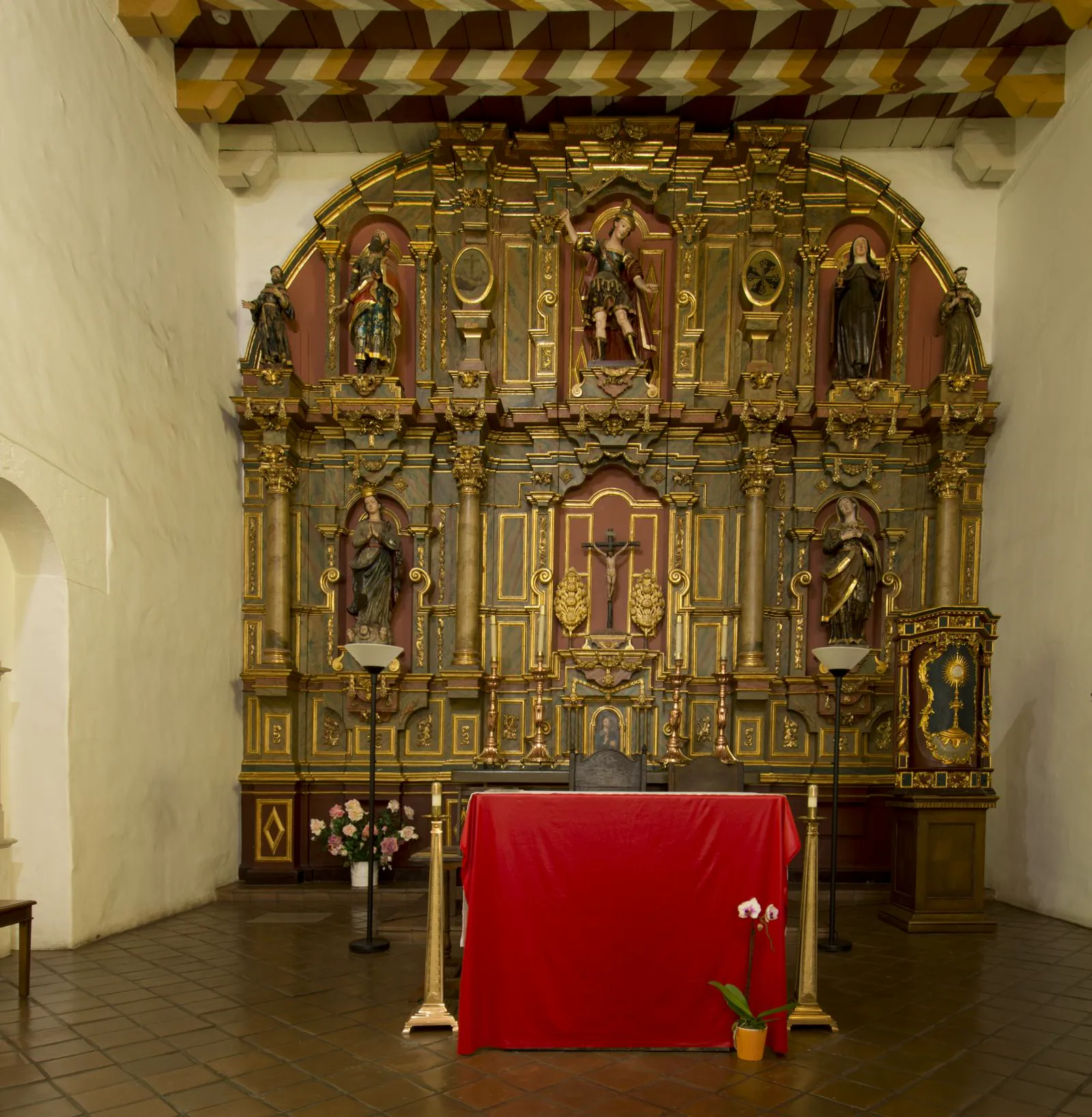 A gilded and painted wooden altarpiece with carved saints in niches and a crucifix at its center rises behind an altar draped in red cloth, under a ceiling of painted beams in red, white and gold