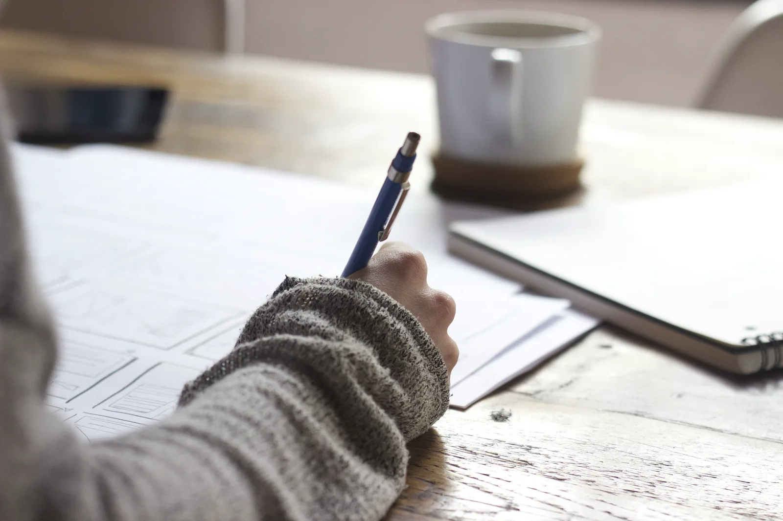 A hand in a gray knit sleeve writes with a blue pen on sheets of paper on a wooden table, beside a white mug and a closed spiral notebook