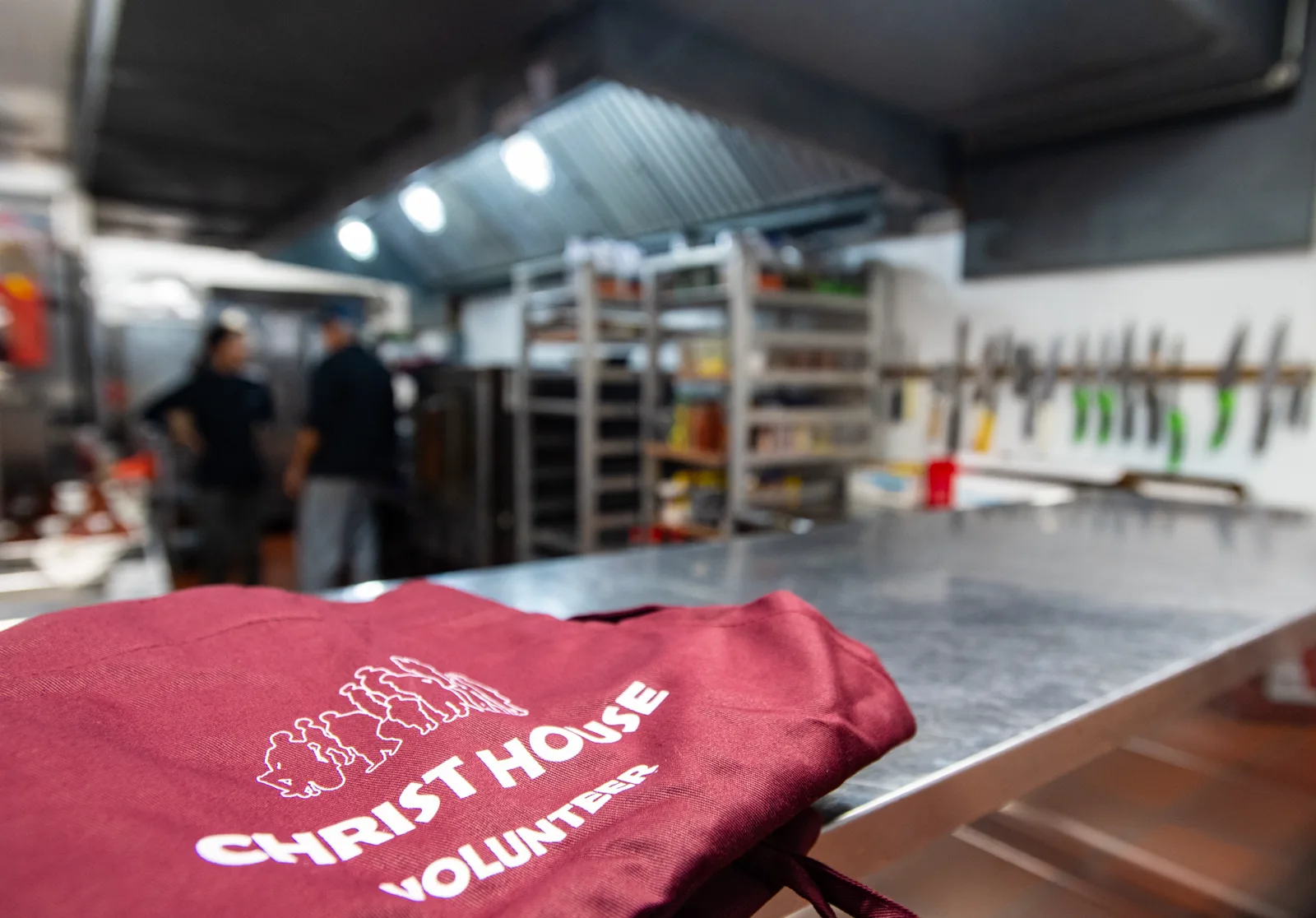 A maroon apron printed with the words Christ House Volunteer lies folded on a steel counter in a commercial kitchen, with two blurred kitchen workers seen from behind near shelving