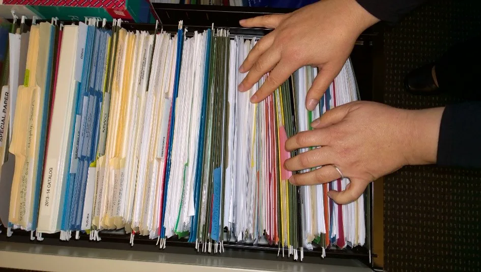 Two hands part a tightly packed drawer of hanging file folders in many colors, with handwritten labels on the tabs