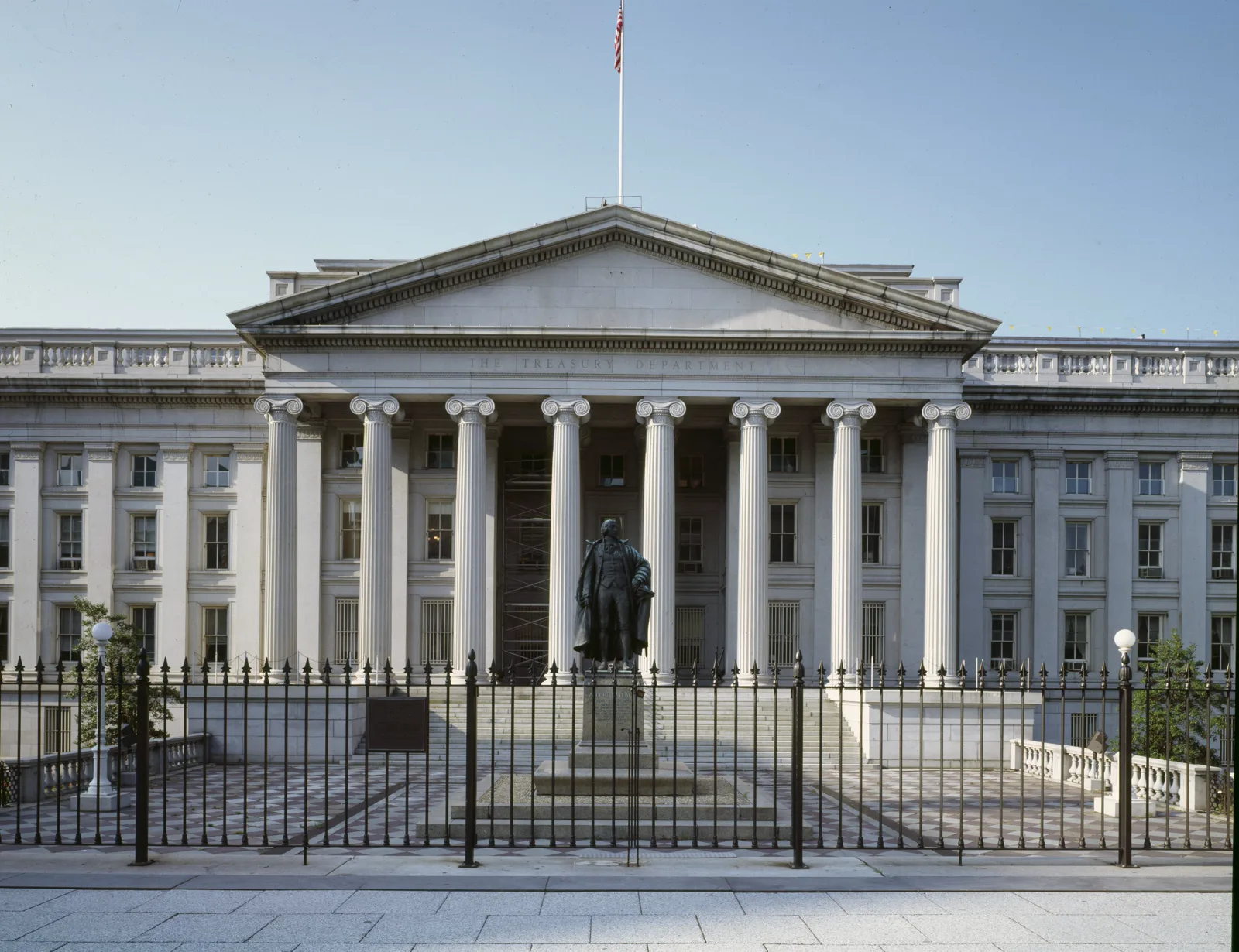 The columned portico of the Treasury Building with The Treasury Department carved above the columns, an American flag on the roof, and a bronze statue on a stone pedestal behind an iron fence in front of the steps