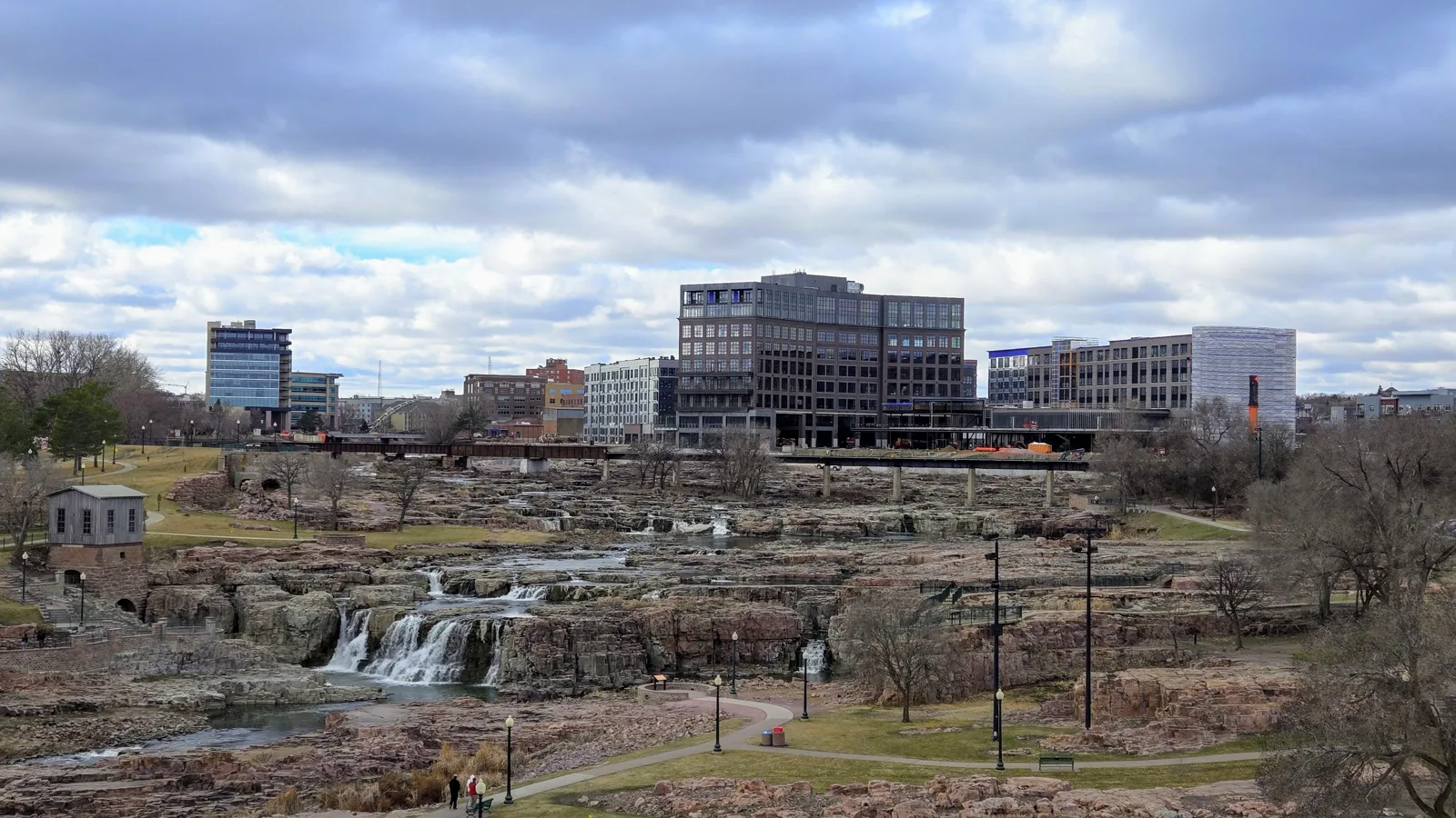 Low waterfalls spill over terraces of pink quartzite in a park with paths, lampposts and bare trees, below a rail bridge and a row of new office and apartment buildings under a cloudy sky