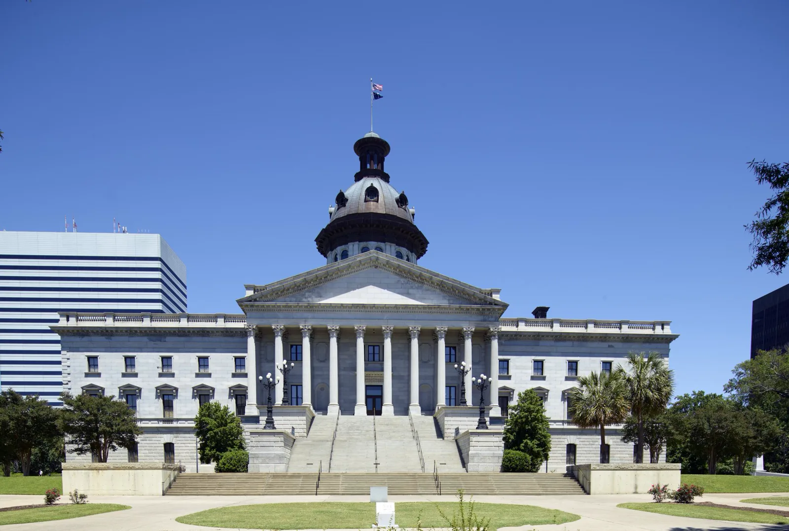 The gray granite South Carolina State House with a copper dome, a columned portico and a wide flight of steps, under a clear blue sky, with palmettos and a tall office building beside it