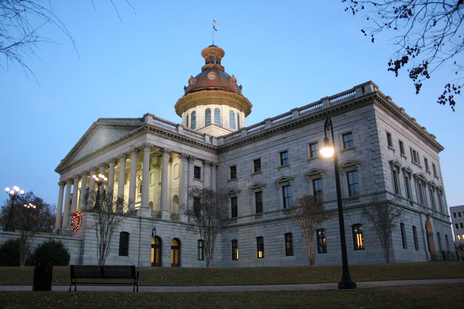 The gray granite South Carolina State House with a copper dome and a columned portico at dusk, its lamps lit, with bare trees and an empty bench on the lawn in front