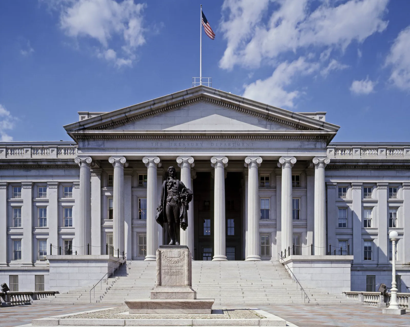 The columned portico of the Treasury Building under a blue sky with an American flag on the roof, and a bronze statue of Alexander Hamilton on a stone pedestal in front of the steps