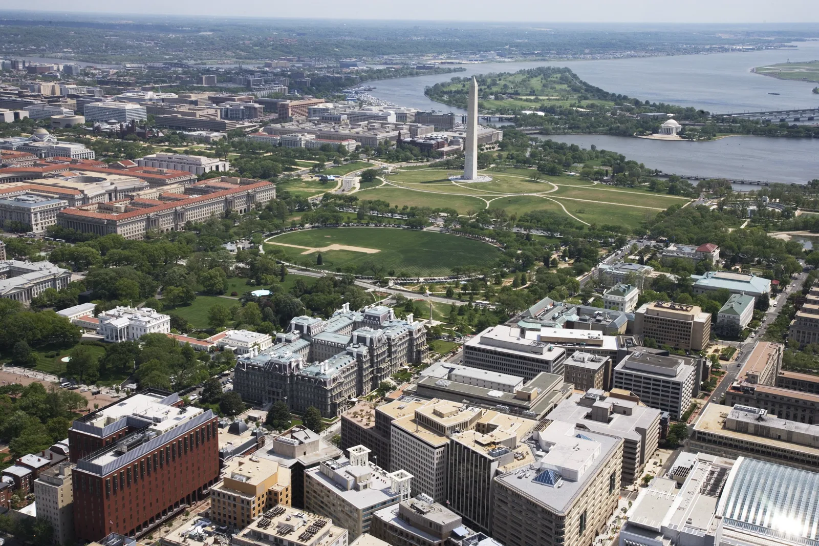 An aerial view of central Washington: the White House among trees at left, a large gray building with mansard roofs beside it, the Ellipse lawn and the Washington Monument beyond, and the Potomac River with a domed memorial in the distance