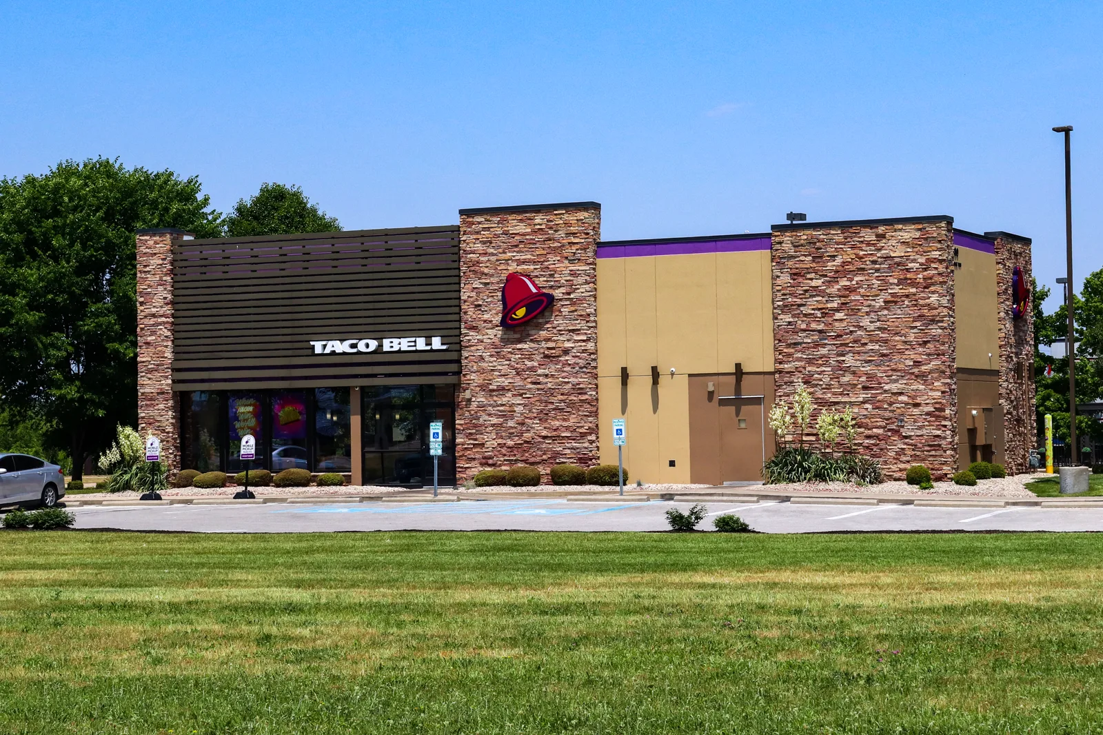 A single-story Taco Bell restaurant with stone-clad walls, a dark slatted front with the Taco Bell name and a bell logo, behind a parking lot and a wide mown lawn under a clear blue sky