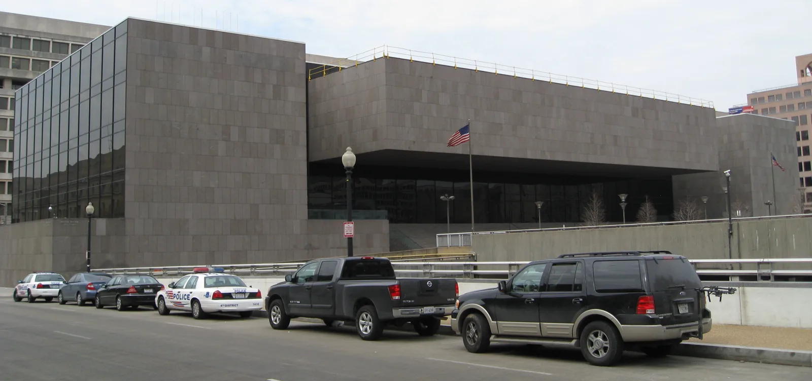 A dark granite building with a long upper block over a recessed glass ground floor and an American flag on a pole in front, with cars parked along the street below
