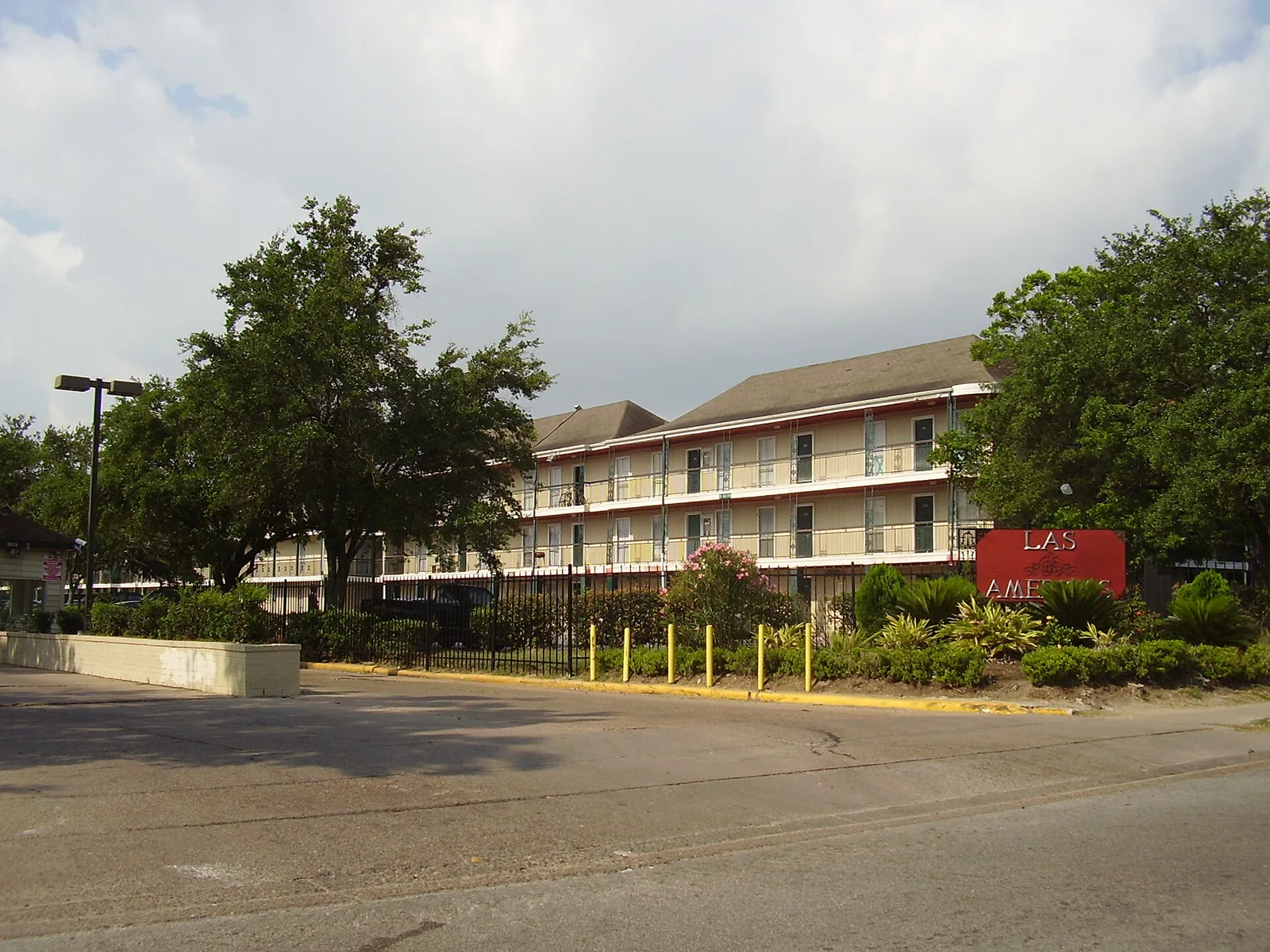 A three story apartment building with balconies behind a black iron fence and trees, with a red sign reading Las Americas at the corner of the lot