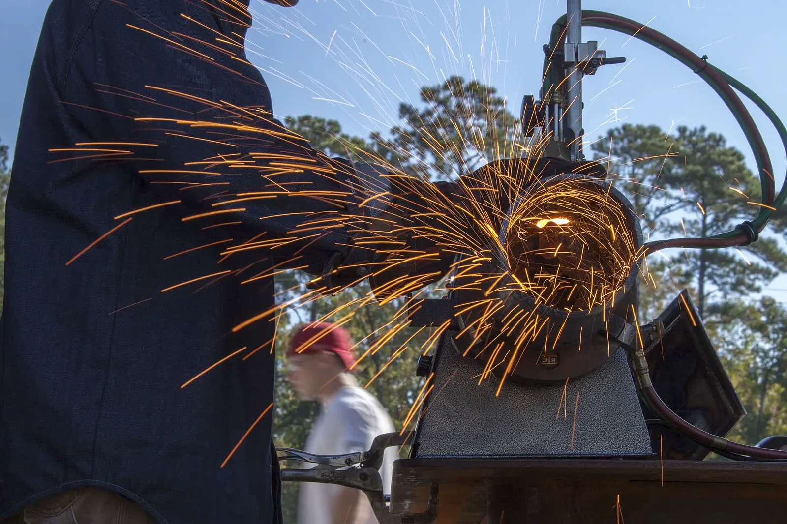 Sparks fly from an automatic welding machine as a man in a dark work shirt operates it under a metal canopy, with pine trees and a passerby blurred in the background