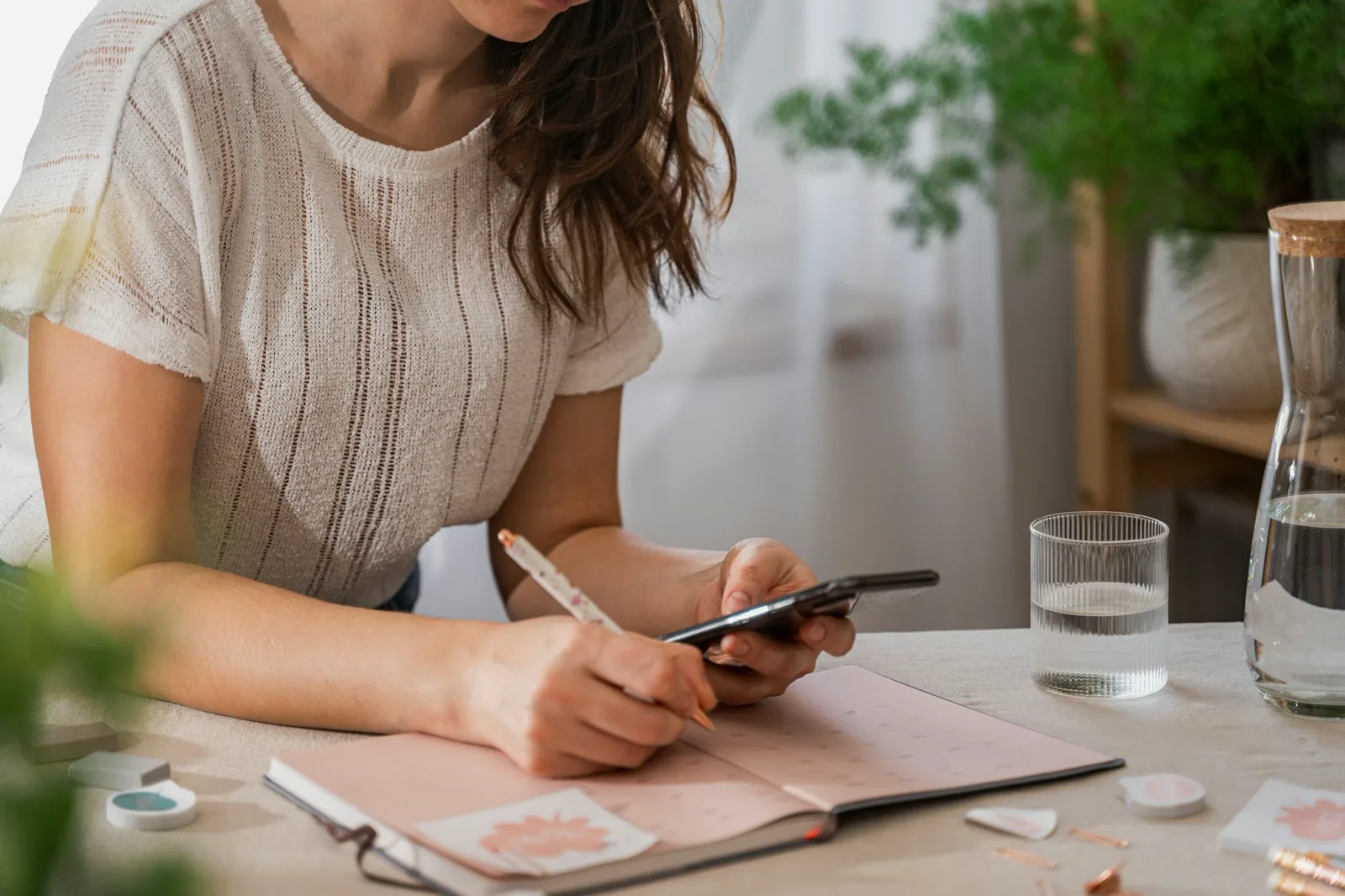 A woman in a white knit top, her face out of the frame, writes in a pink weekly planner at a table while holding a phone, beside a glass and a carafe of water