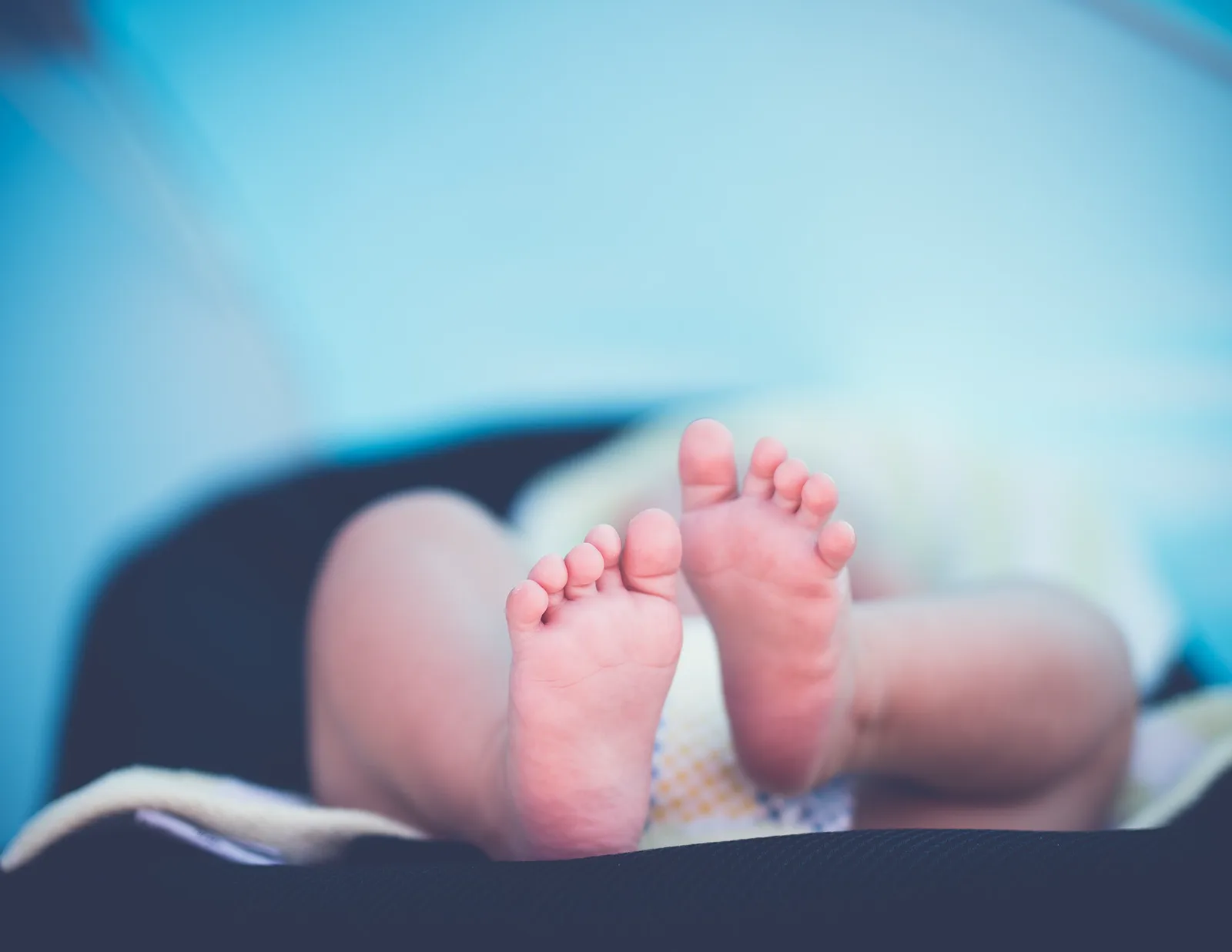 A newborn baby's bare feet in sharp focus, the legs blurred behind them, lying on a dark padded seat against a pale blue background