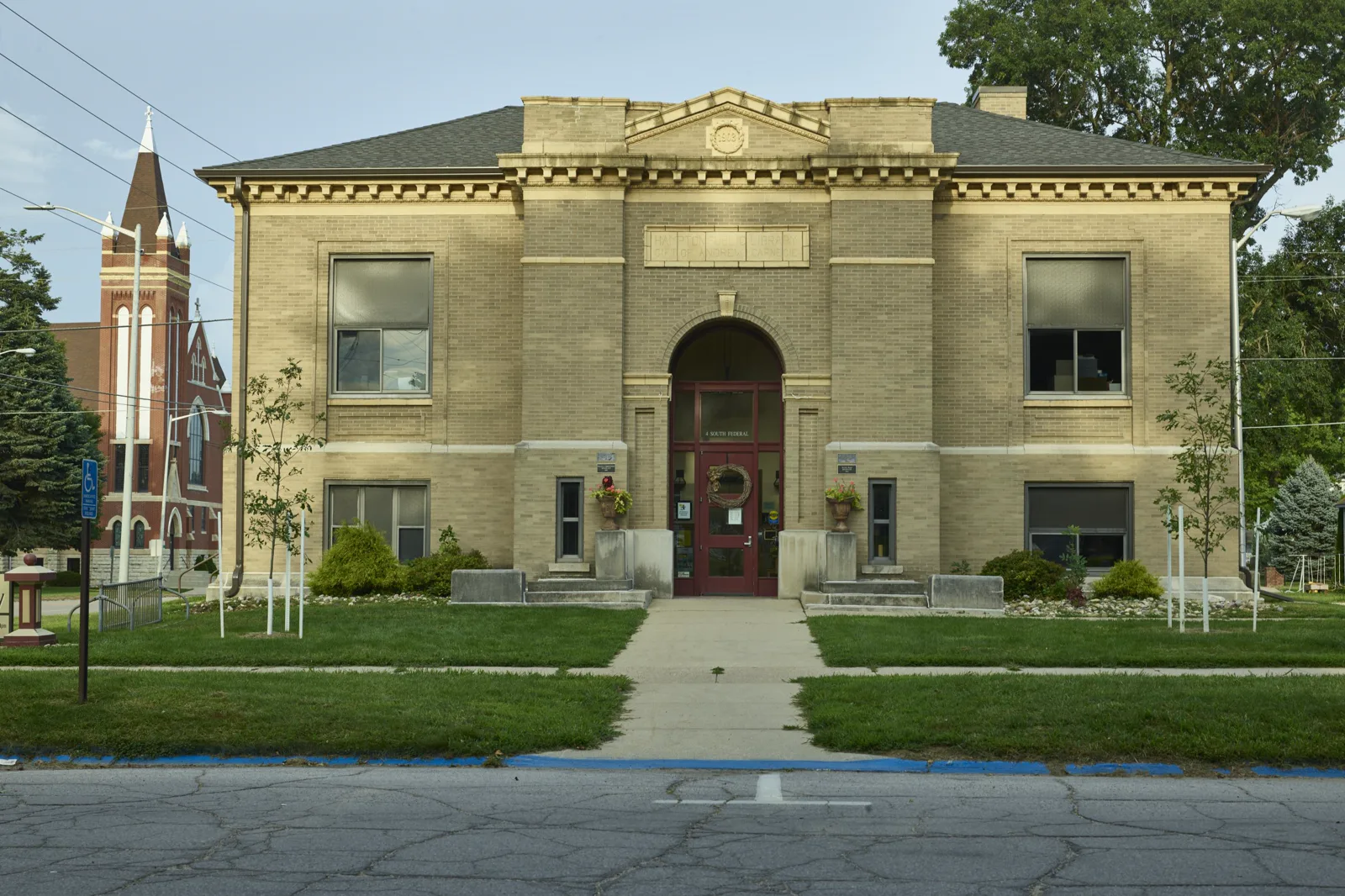 A two-story tan brick library building with a low hipped roof and an arched entrance with a red door and a wreath, young trees on the lawn in front and a red brick church spire at left, seen across a street