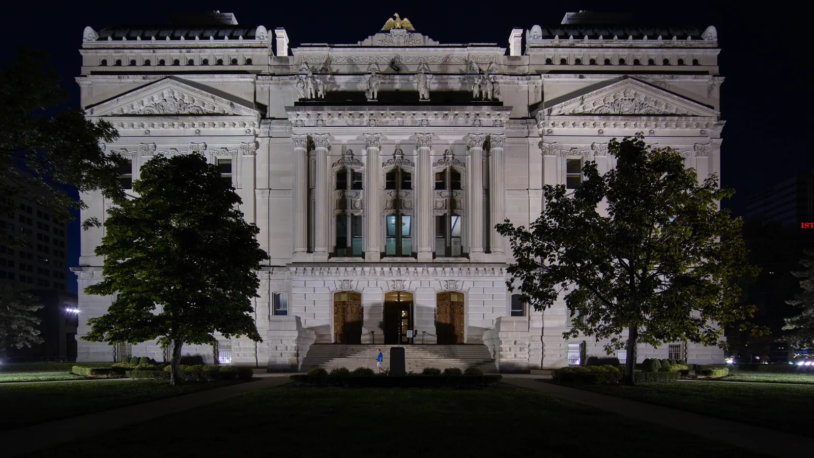 The pale stone facade of the Indiana Statehouse lit up at night, with tall columns, a row of statues and a gilded eagle at the top, framed by two trees on a dark lawn, and one person walking across the front steps